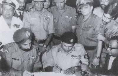 Lt Gen Niazi signing the Instrument of Surrender under the gaze of Lt Gen Aurora . Standing immediately behind from L to R: Vice Admiral Krishnan, Air Marshal Dewan, Lt Gen Sagat Singh, Maj Gen JFR Jacob (with Flt Lt Krishnamurthy peering over his shoulder). Veteran newscaster, Surojit Sen of All India Radio, is seen holding a microphone on the right. Photo: Wikimedia Commons